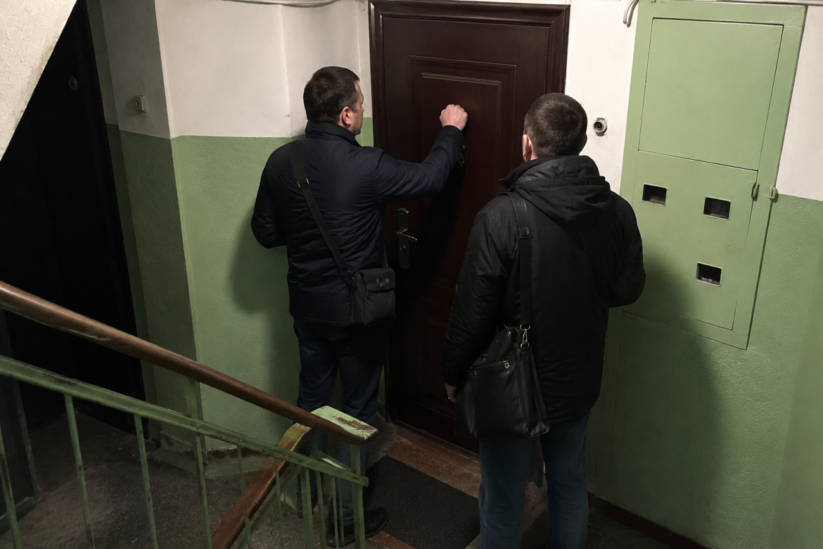 Двоє чоловіків у темному одязі стукають у двері квартири в під'їзді